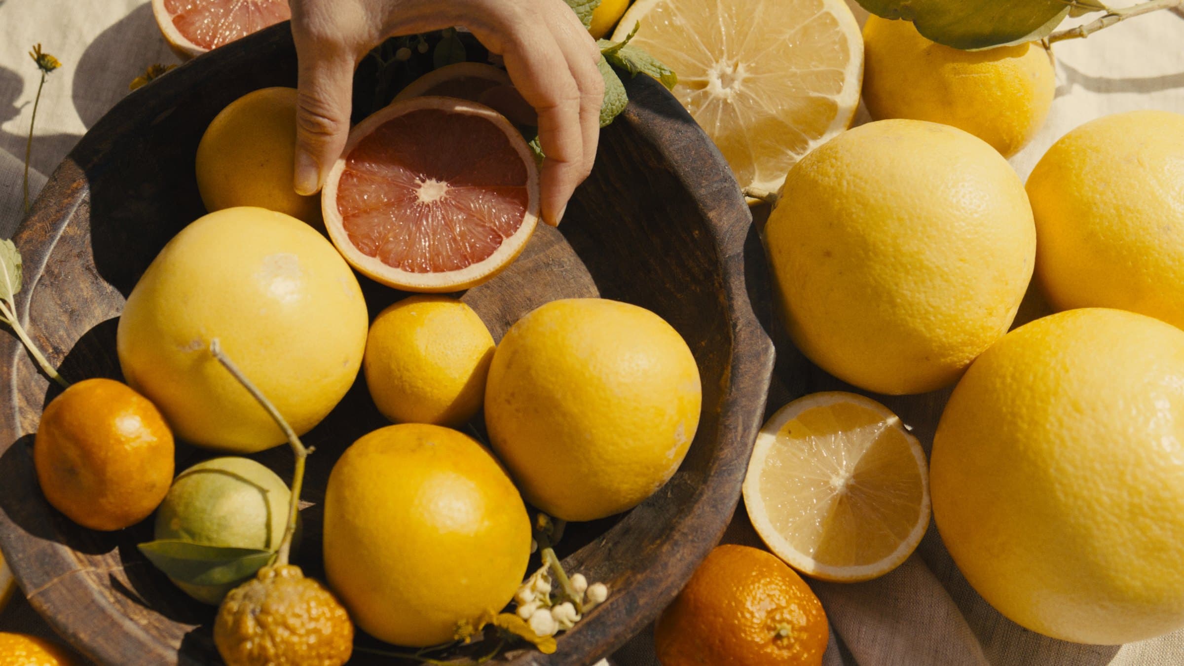 A hand reaches into a wooden bowl of lemons, grapefruit and mandarins on a linen cloth, holding a halved pink grapefruit in sunlight.