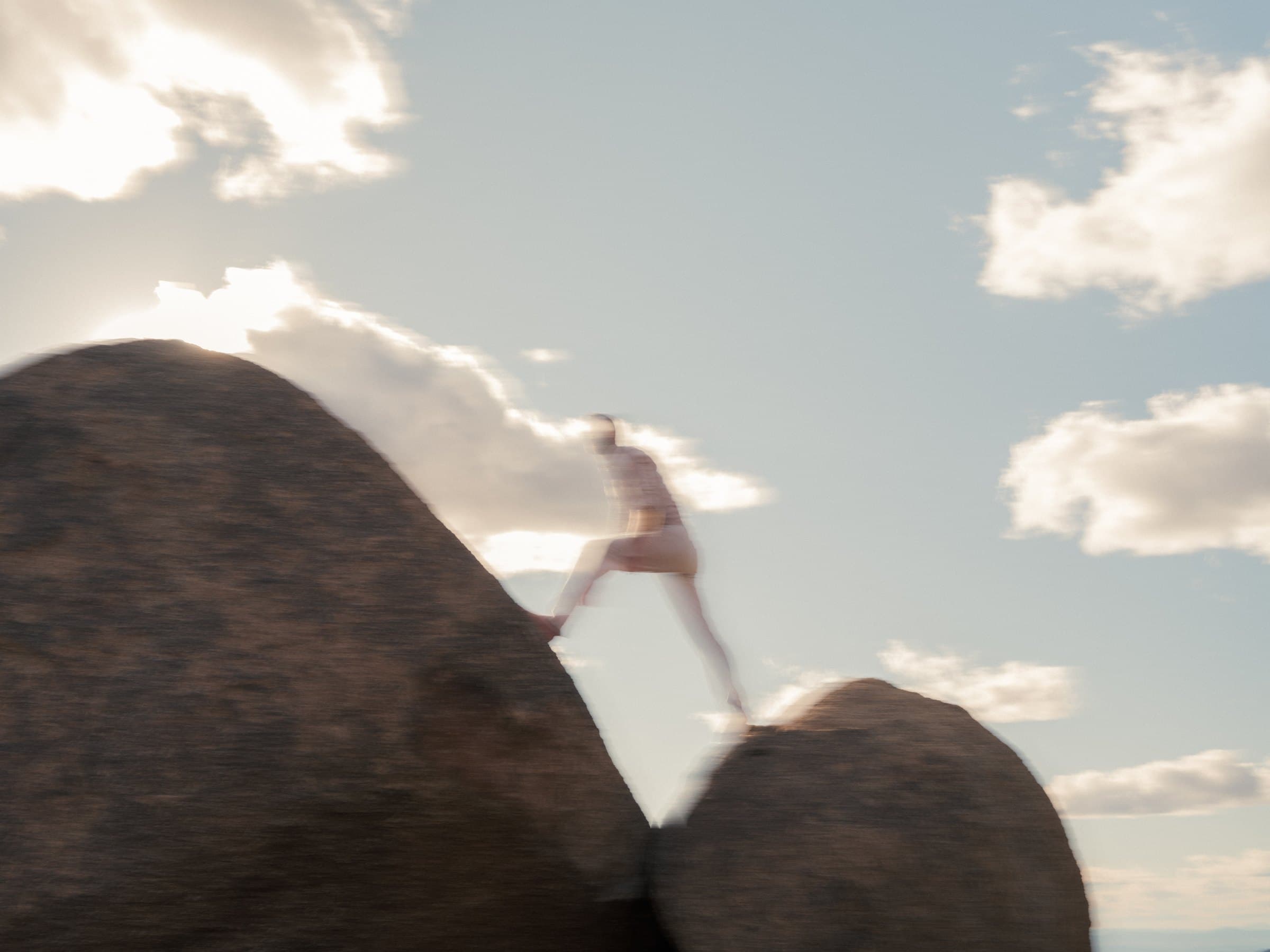 A blurred figure in light clothing steps across the gap between two large rounded boulders, photographed against a bright sky with scattered clouds.