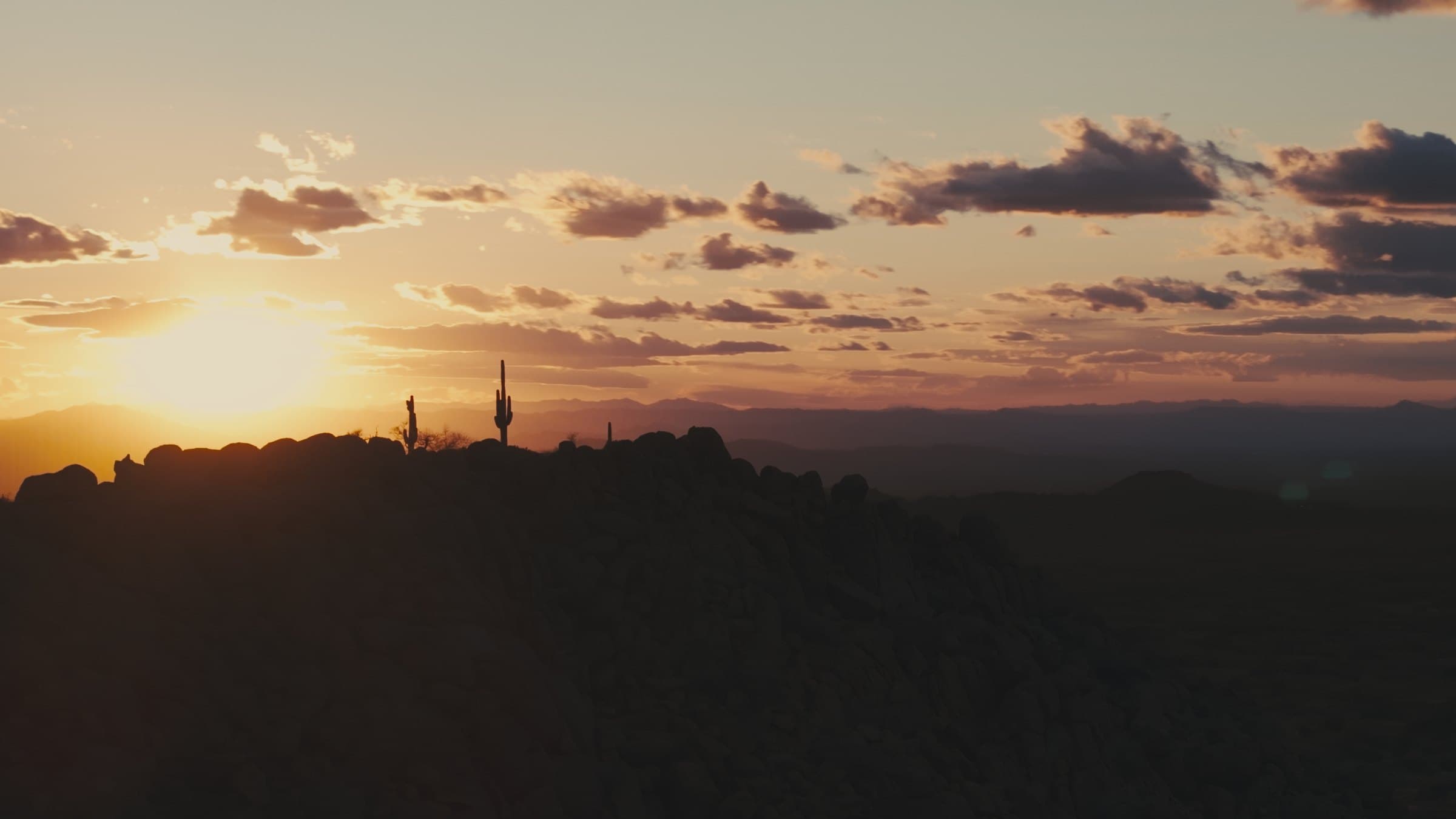 A rocky desert ridge in silhouette at sunset, with saguaro cactus on the skyline and distant mountain ranges under a cloud-streaked orange sky.