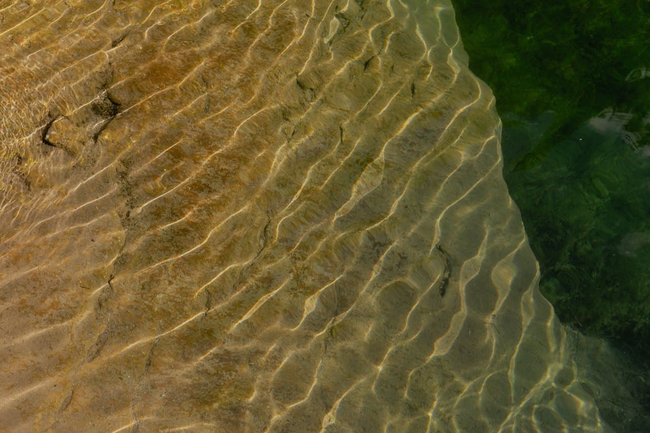 Looking straight down at clear shallow water over rippled sand, with bands of sunlight on the bottom and darker green deep water along the right side.