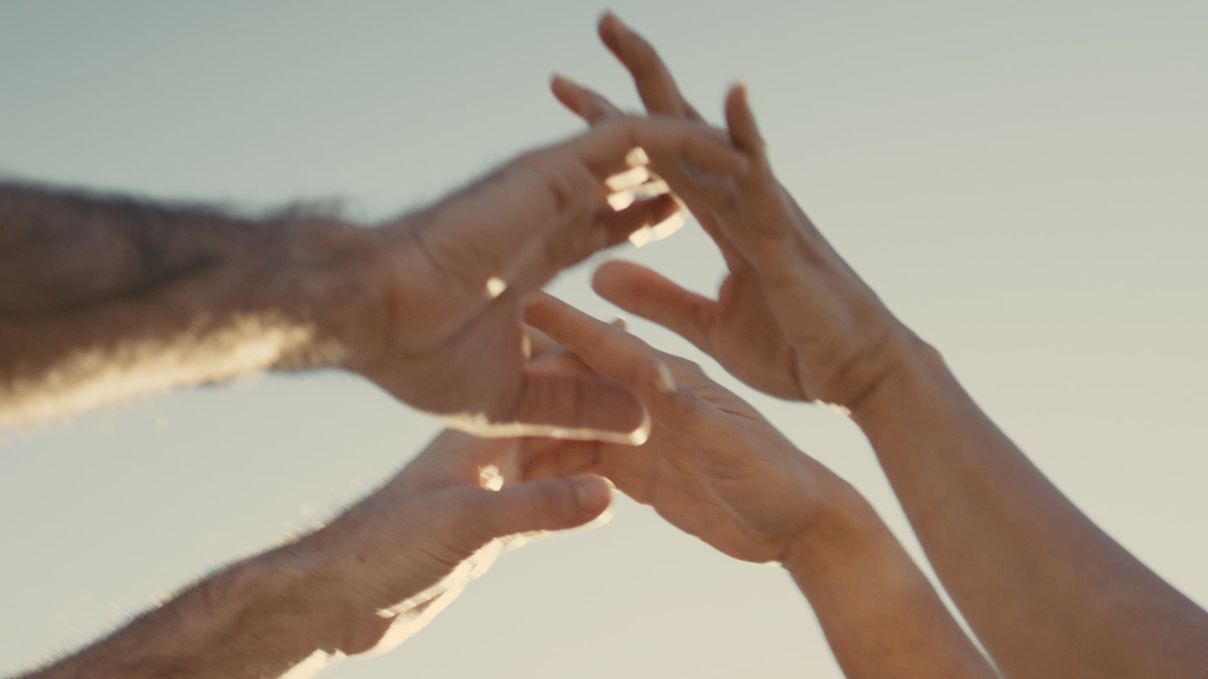 Two pairs of hands reaching toward one another with fingers almost touching, photographed from below against a pale sky and lit from behind.