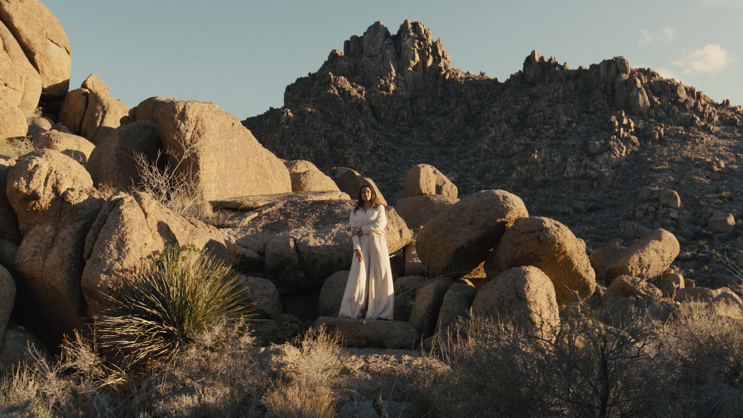 A woman in cream clothing stands on a large granite boulder in a desert boulder field, with a rocky ridge and a yucca plant behind her.