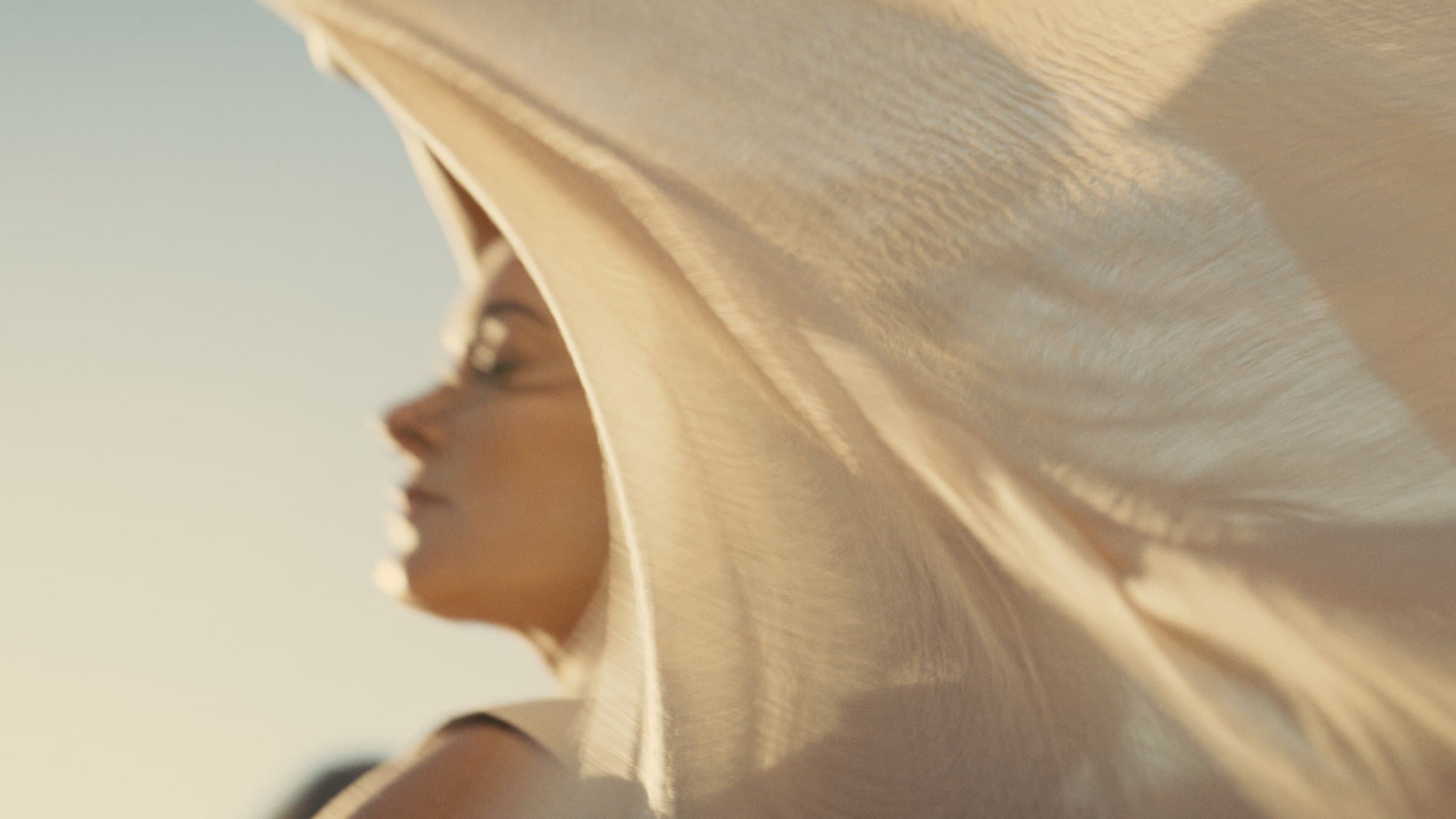 A close side profile of a woman with a cream linen scarf blowing across her head and shoulder, lit from behind against a pale sky.